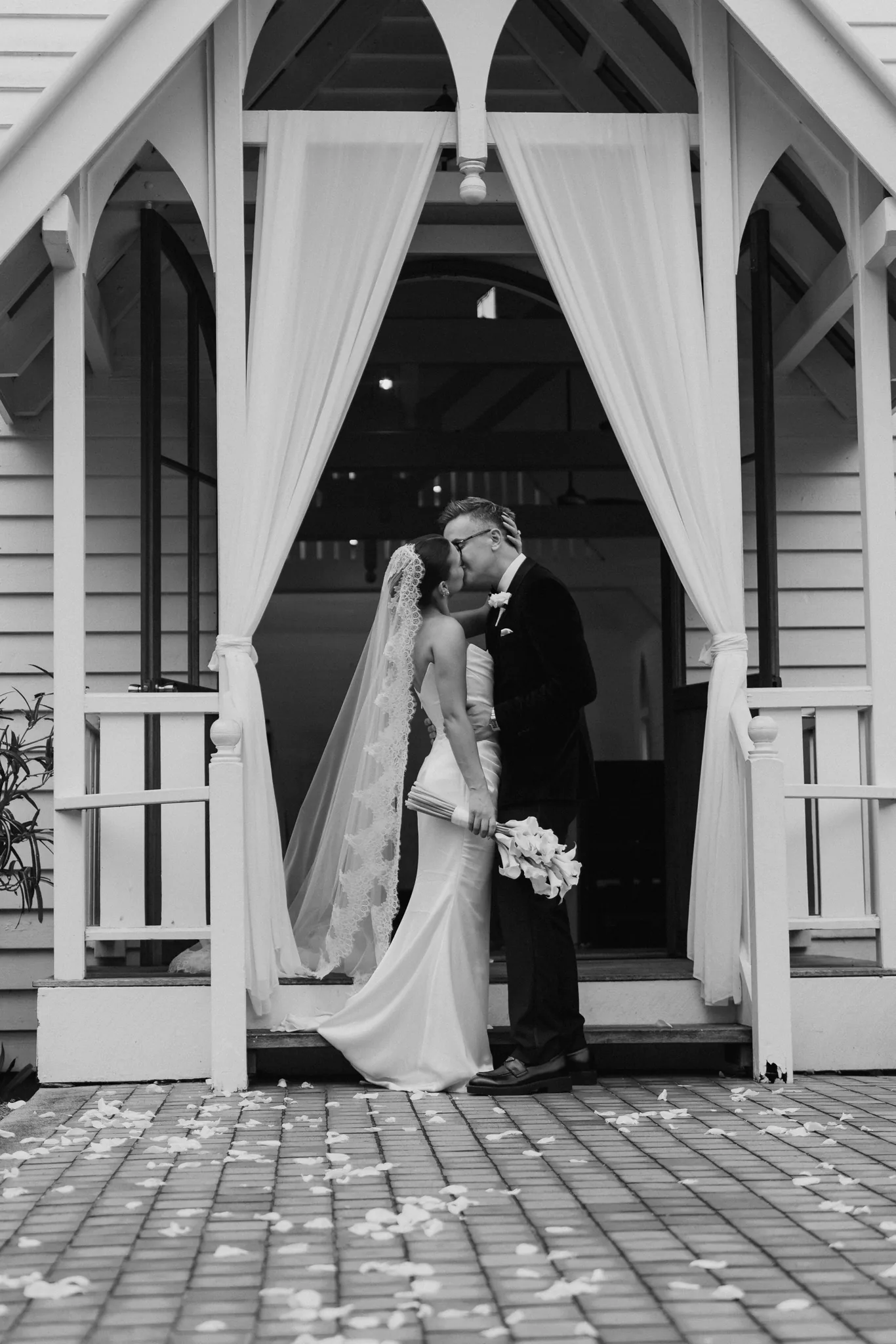The couple shares a kiss in the draped doorway of a white chapel, in black and white