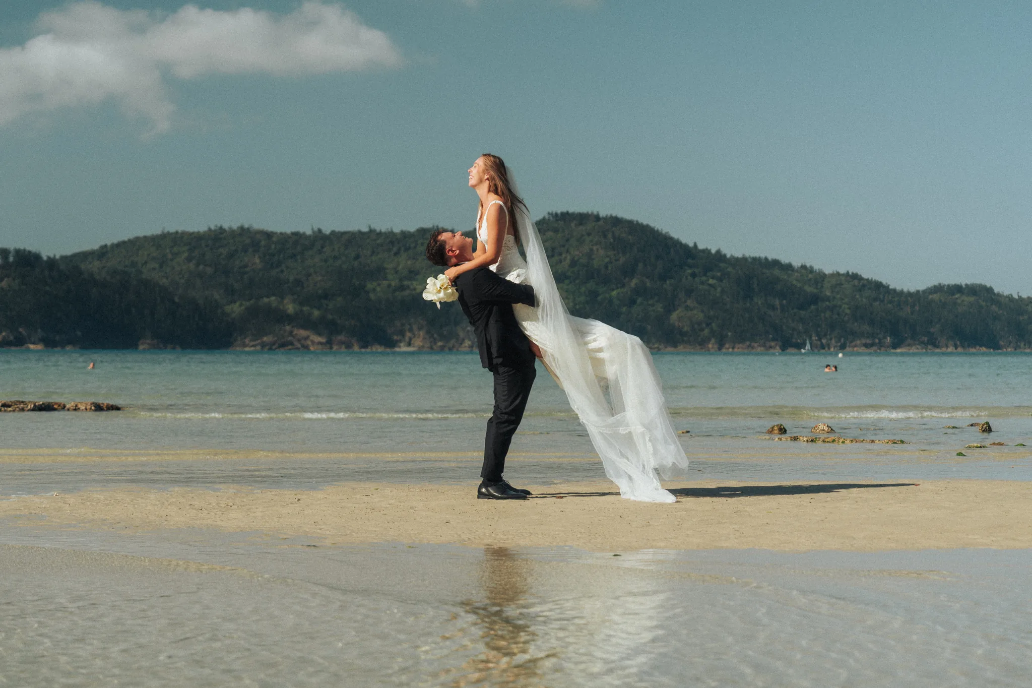 A groom dips the bride for a kiss on wet sand, with mountains beyond