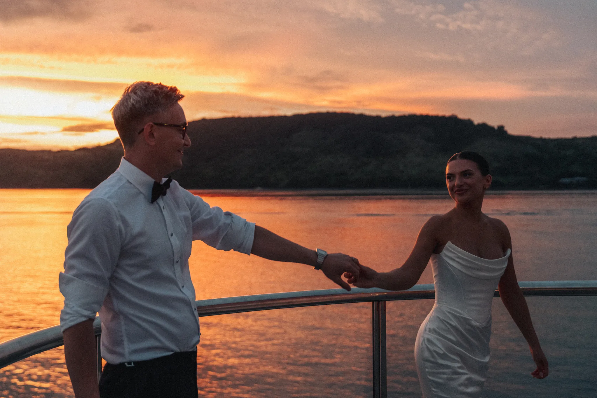 A couple stand at a railing, facing each other against a sunset over the water