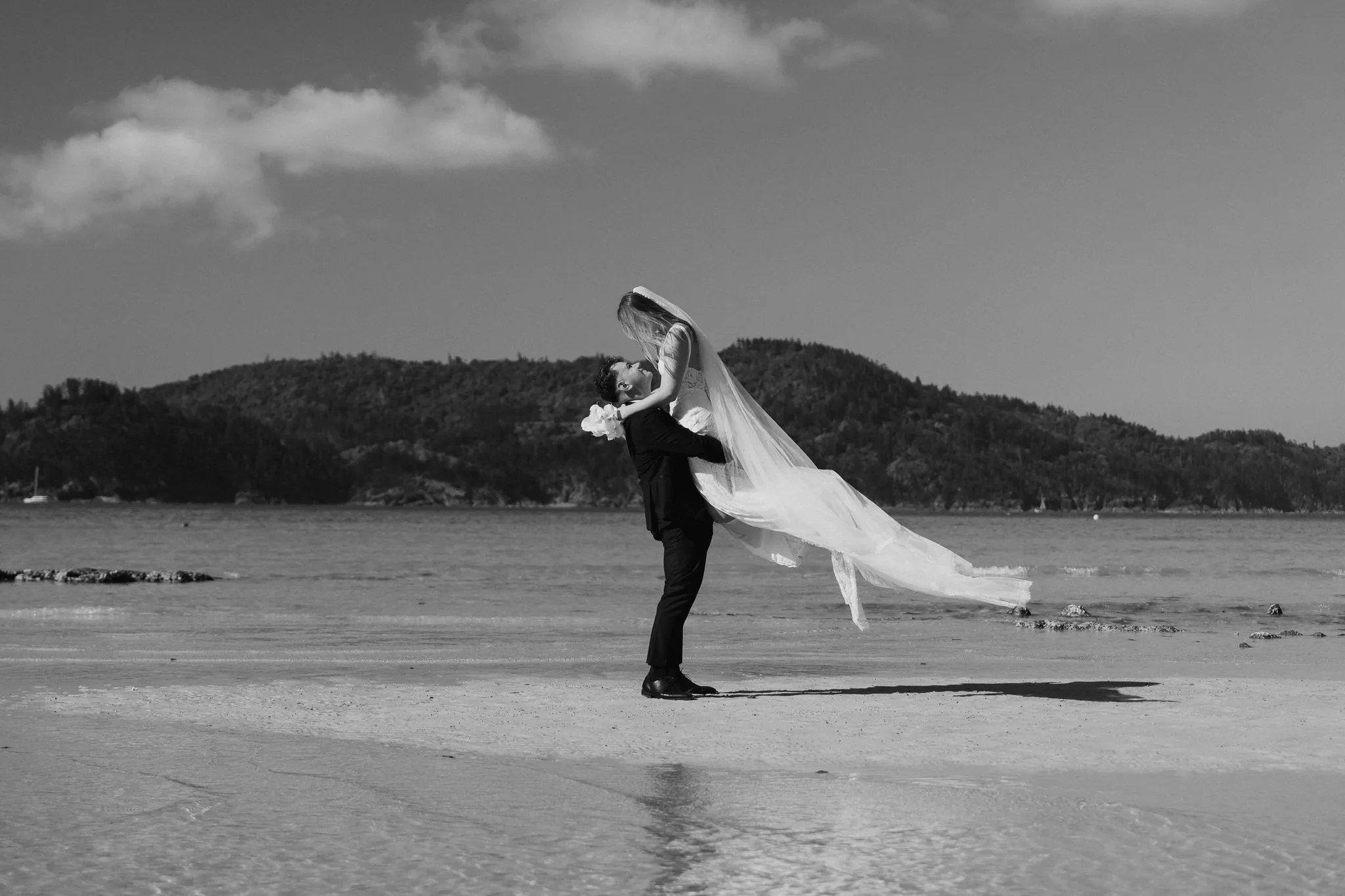 The groom twirls the bride across an open beach, in black and white